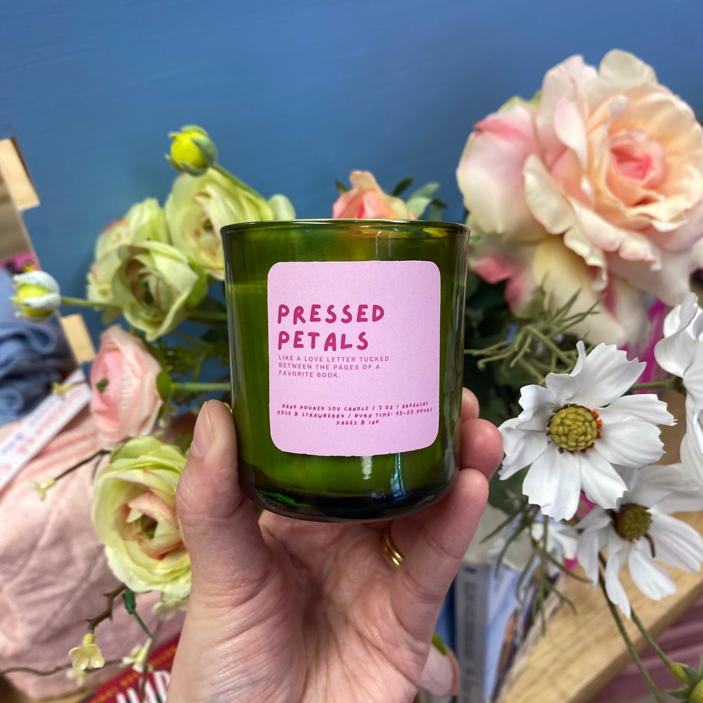 Hand holding a 'Pressed Petals' candle with flowers in the background