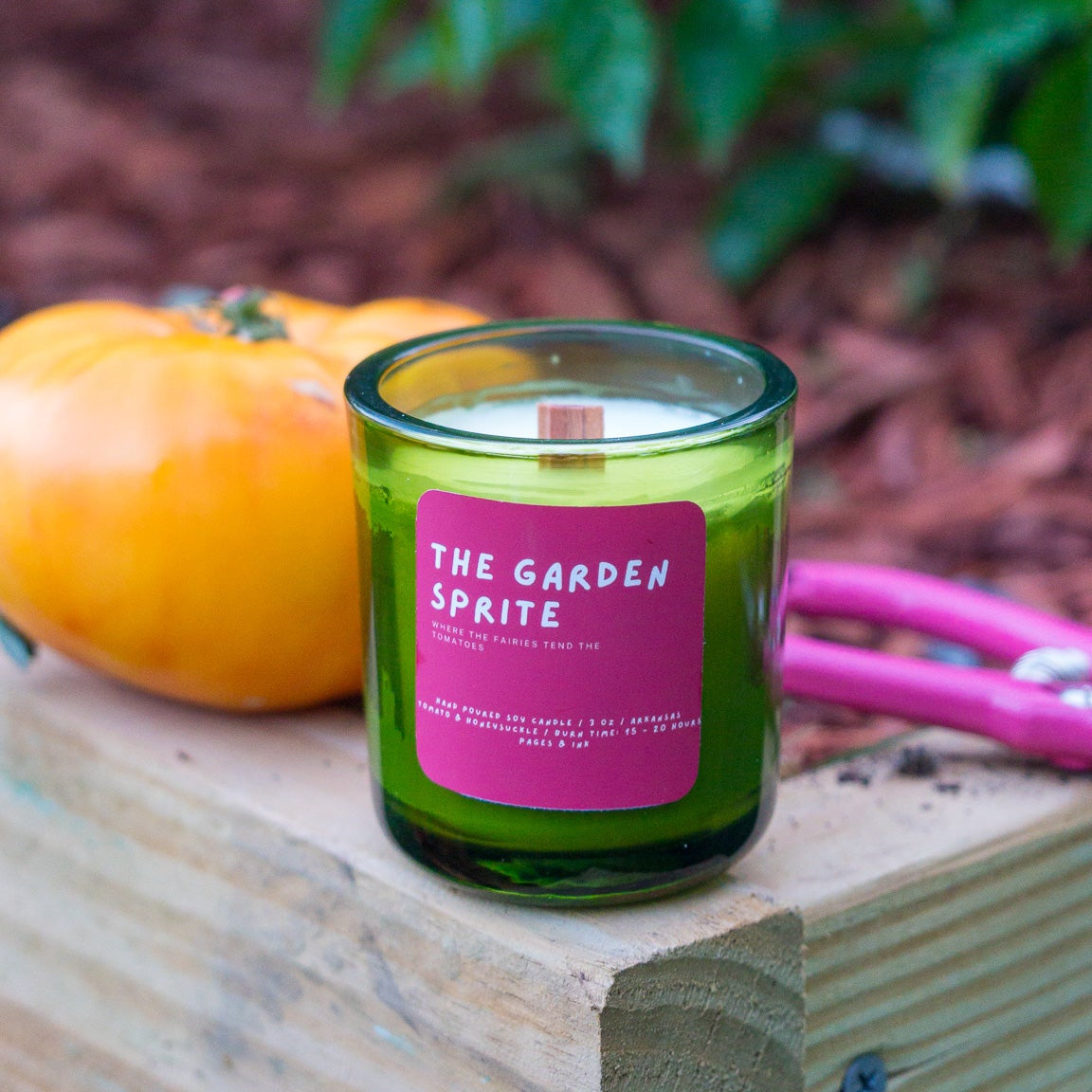 A green glass jar candle with a pink label reading 'THE GARDEN SPITE' sits on a wooden crate outdoors, next to a yellow-orange pumpkin and pink pruning shears, with blurred green foliage in the background.