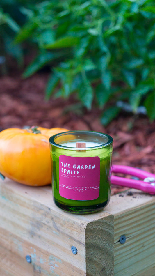 A green glass jar candle with a pink label reading 'THE GARDEN SPITE' sits on a wooden crate outdoors, next to a yellow-orange pumpkin and pink pruning shears, with blurred green foliage in the background.