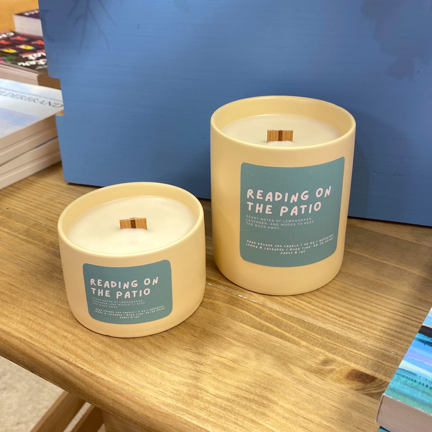 Two candles labeled 'Reading on the Patio' on a wooden surface with books and a blue box in the background.