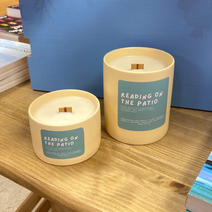 Two candles labeled 'Reading on the Patio' on a wooden surface with books and a blue box in the background.