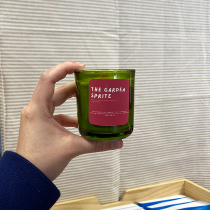 Hand holding a green candle labeled 'The Garden Sprite' in a bookstore setting with books in the background.