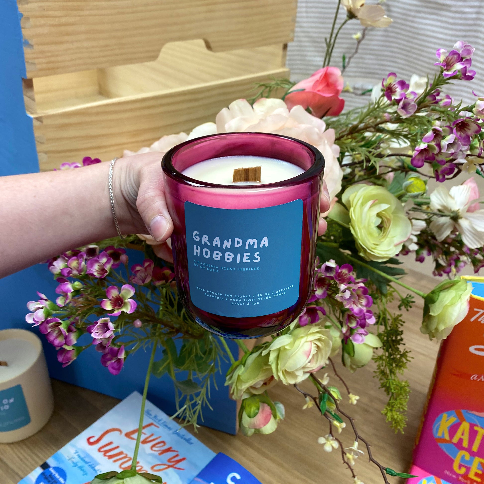 Hand holding a candle labeled 'Grandma Hobbies' with flowers and books in the background