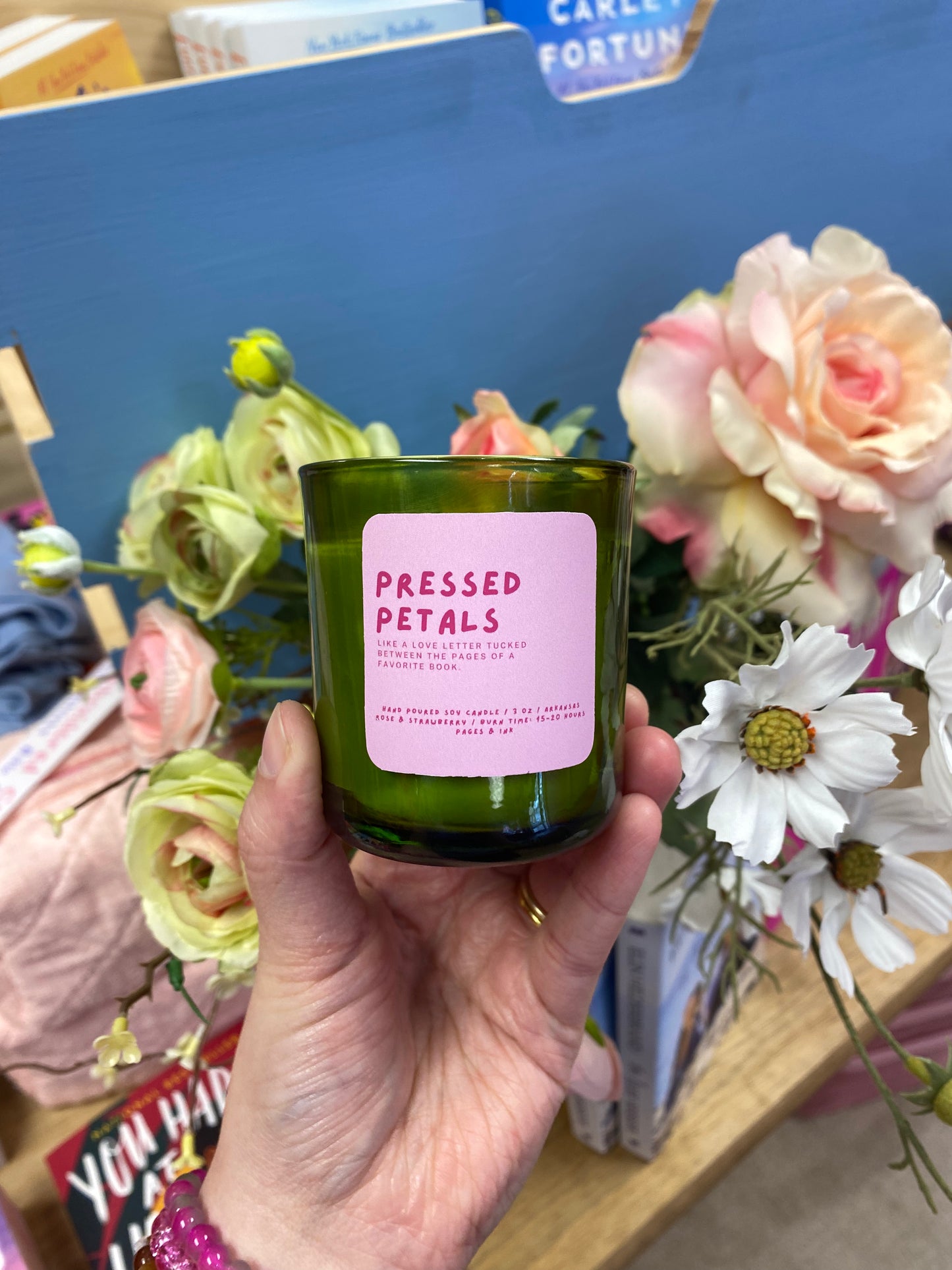 Hand holding a 'Pressed Petals' candle with flowers in the background